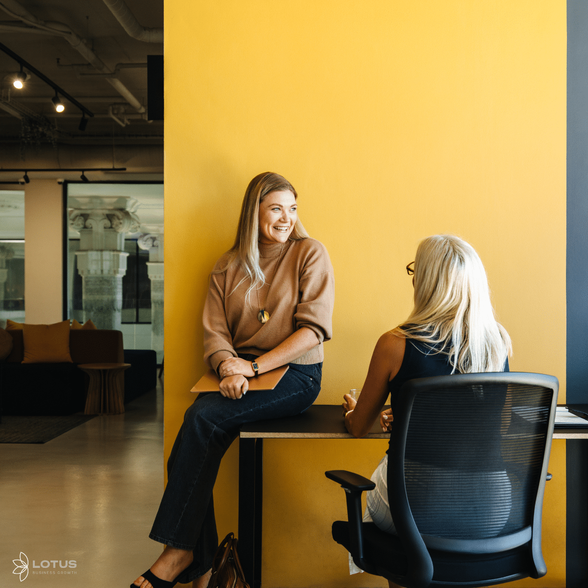 Two women having a casual conversation in a modern office beside a bright yellow wall.
